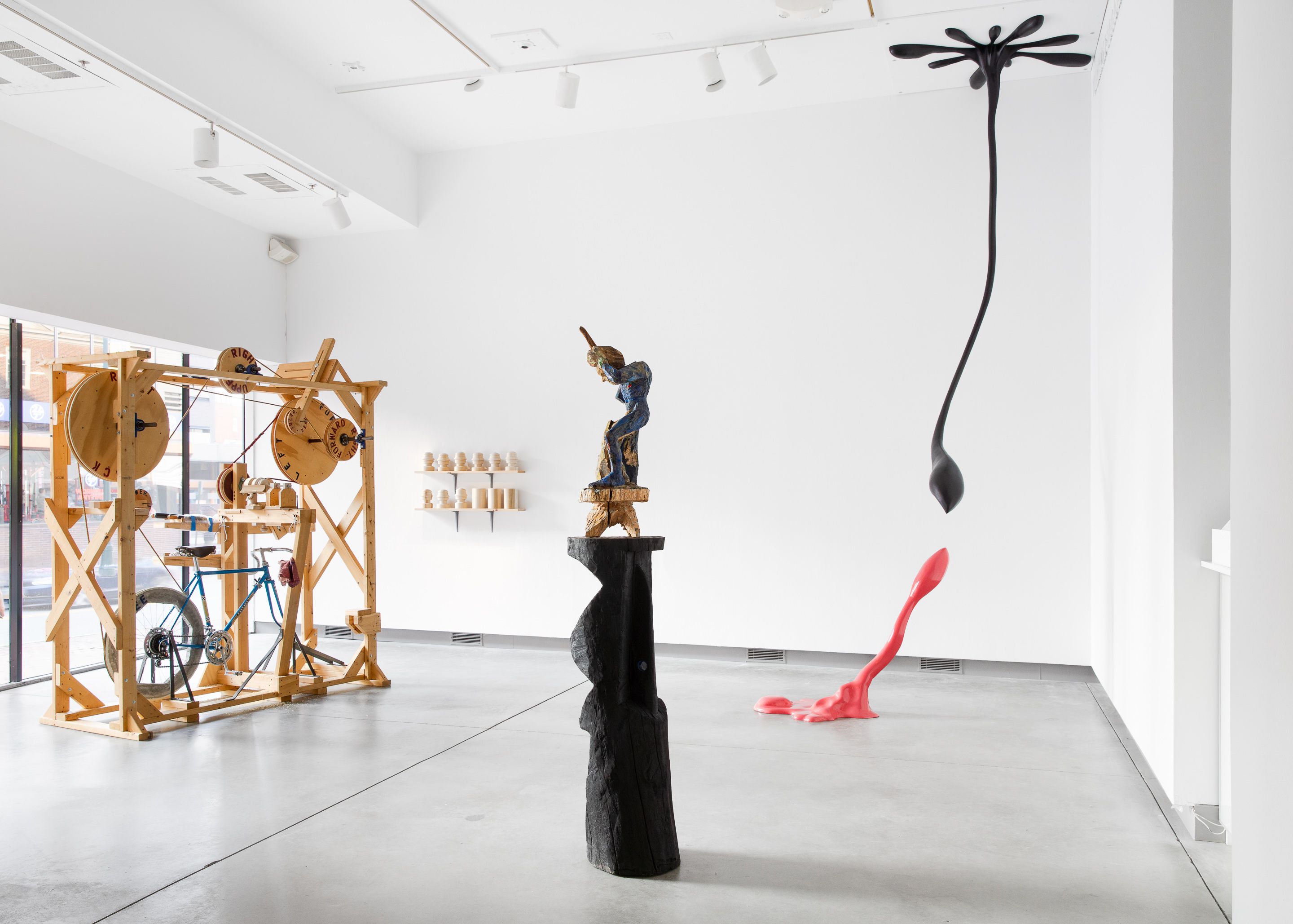 Installation view of three sculptures, the left a bike-powered kinetic wooden frame, the middle a figure seated on a pedestal, the right two liquid-like forms reaching to each other.