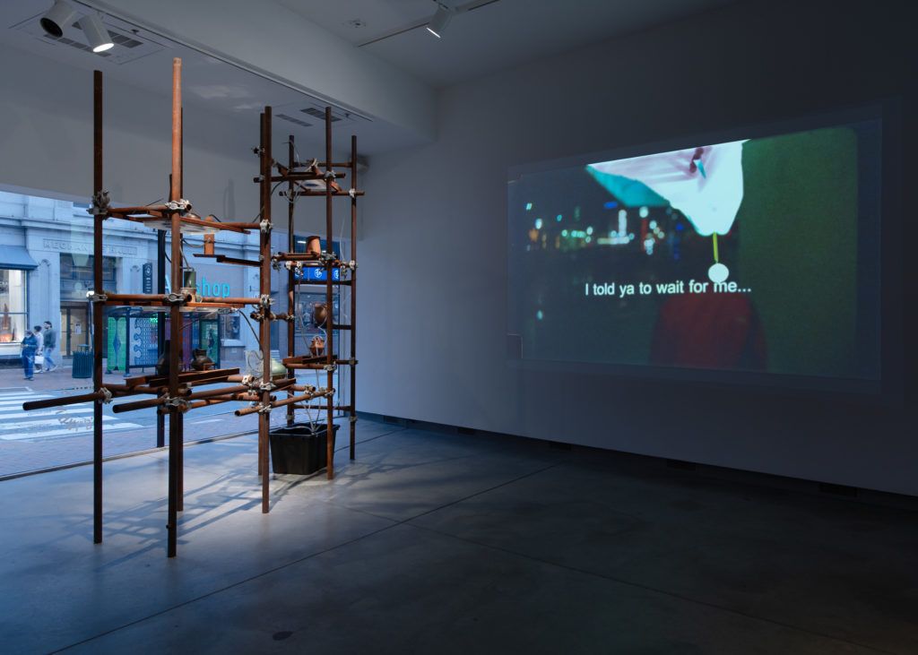 A shelf-like sculpture with water tubes next to a projection with the text, "I told ya to wait for me..."