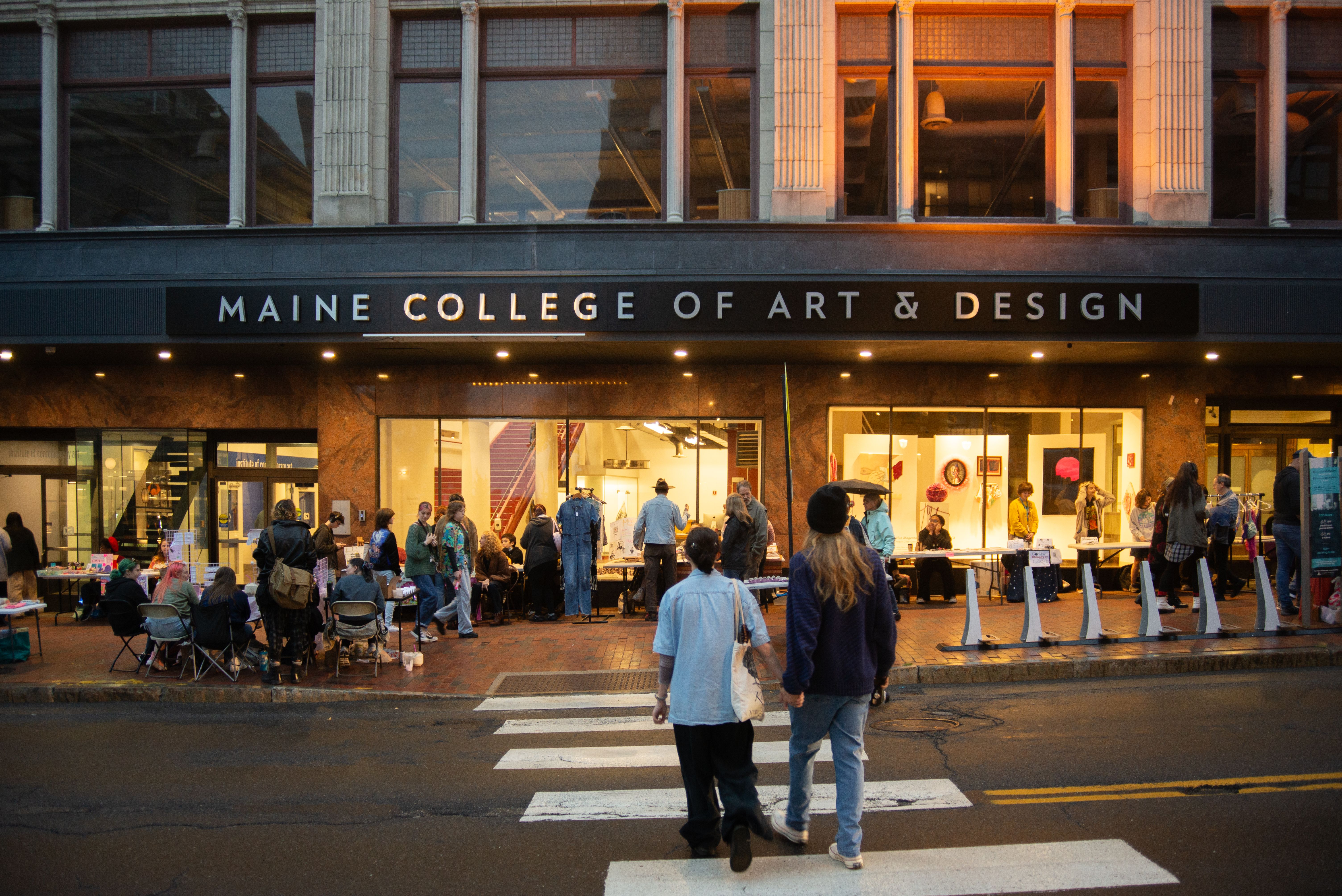 Two people crossing the street towards the Porteous Building where there are many tables set up to sell art.