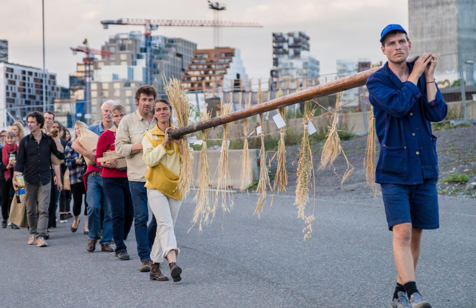 A group of people walking single-file. The first two are carying a large wooden pole tied with dried plants on their shoulders.