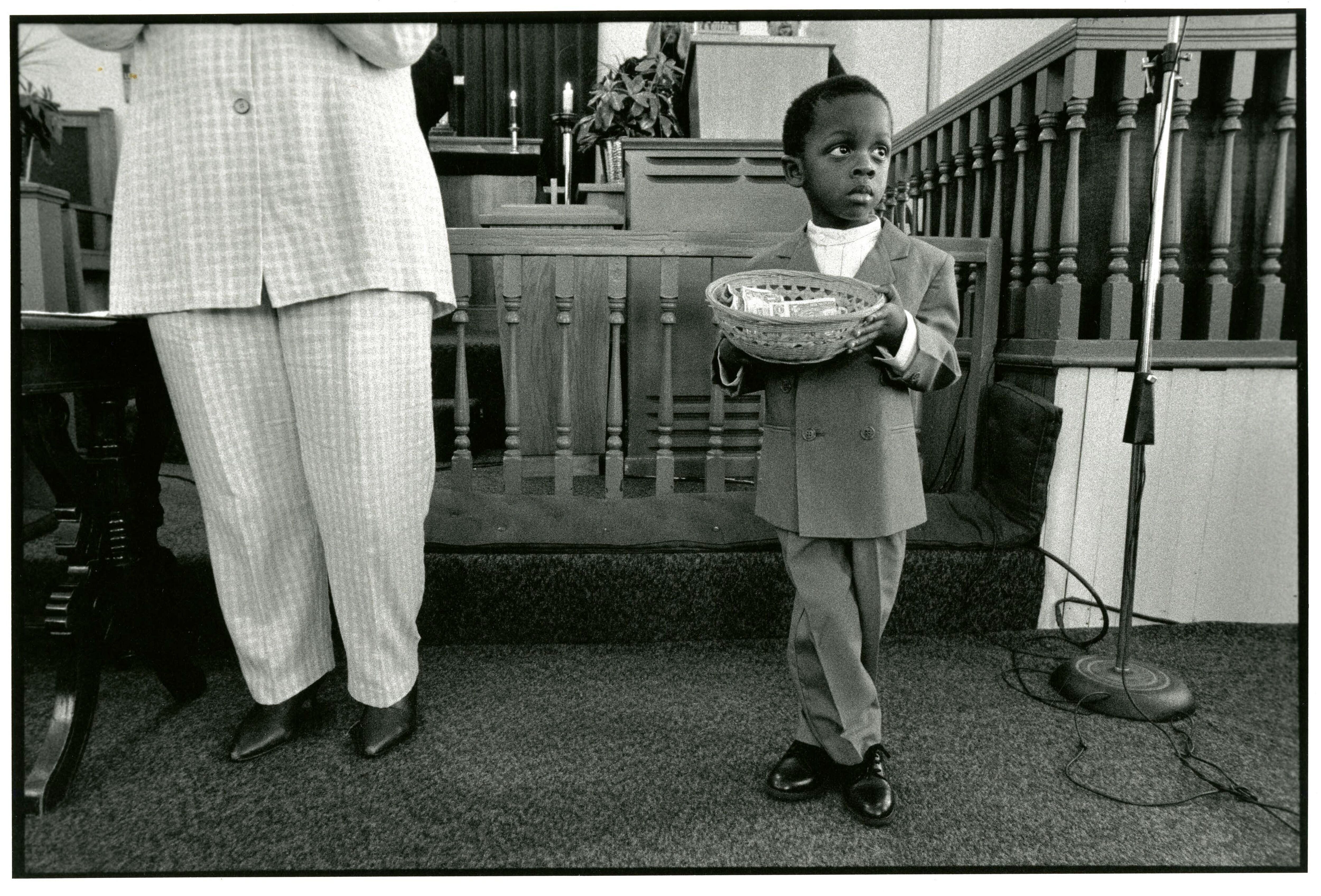 A black and white photograph of a child in a suit holding a donation basket in front of a church alter. Next to him is the lower half of an adult in a white suit.