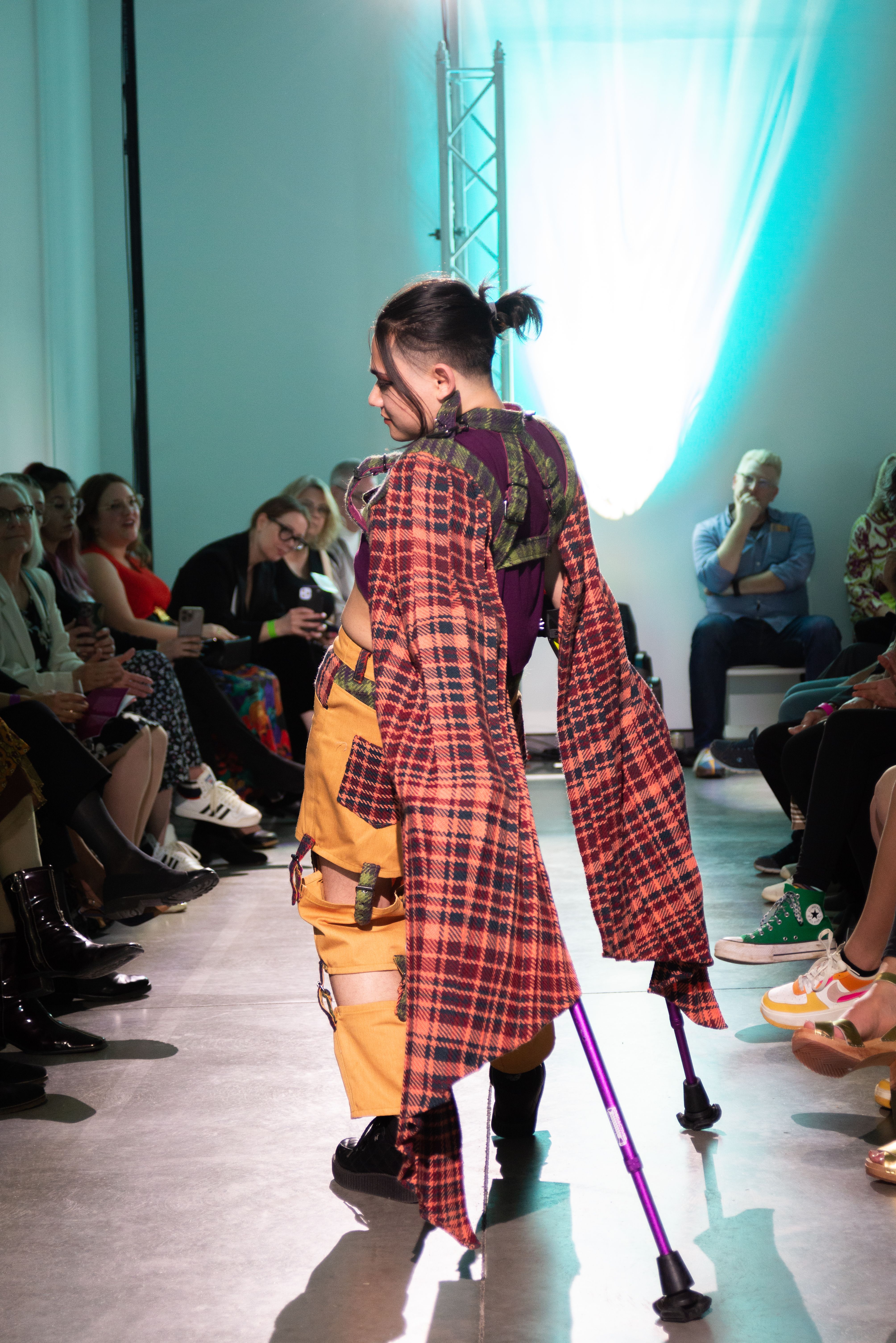 A model on the runway posing with their canes wearing a plaid cape and yellow pants with plaid details.