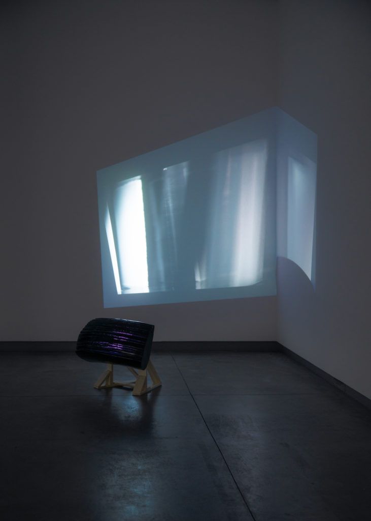 Gallery view of a distorted blue projection above a short sculpture.