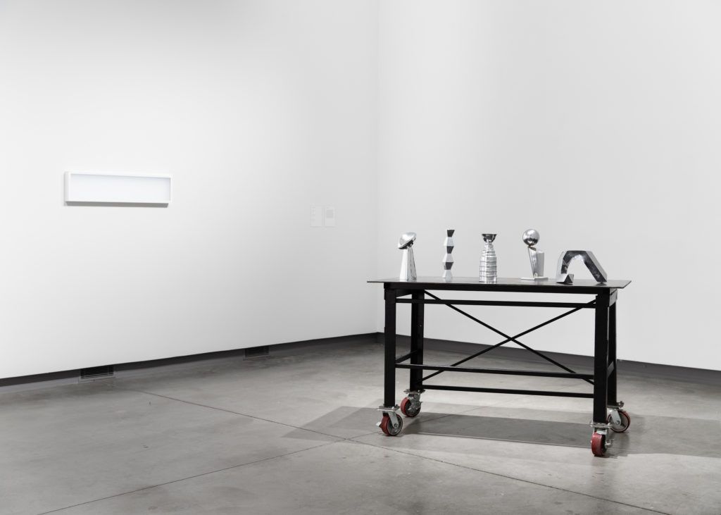 Gallery view of a framed white rectangle piece next to a black cart carrying five metallic structures.