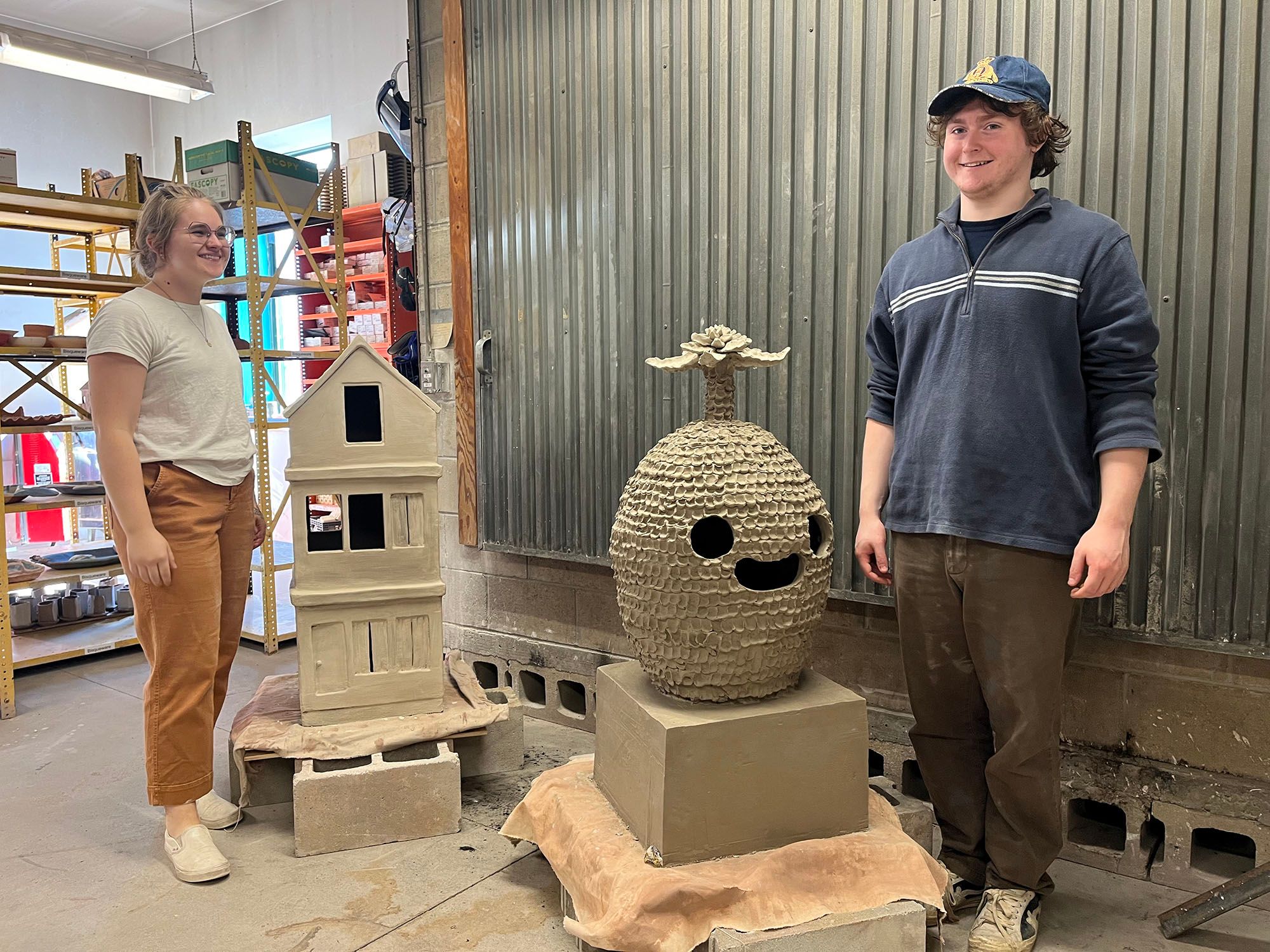 Two students with three foot tall sculptures ready to fire in the kiln. The left is a three-story house, the right is a palm tree-like character.