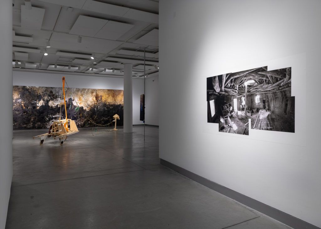 A gallery view of a black and white irregularly shaped photograph, a wooden sculpture, and a large abstract painting.