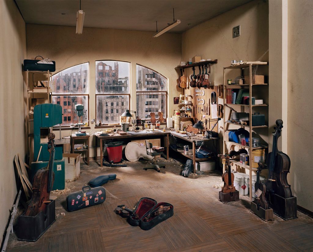 A depiction of a cluttered violin repair shop overlooking decaying buildings.