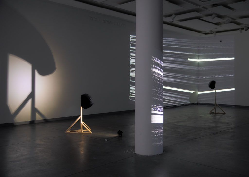 Gallery view of a projection of stripes and two sculptures.