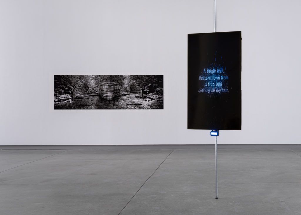 A hanging screen with glowing fading text that reads "A single leaf, flutters down from a tree, and settling on my hair" next to a black and white landscape print.