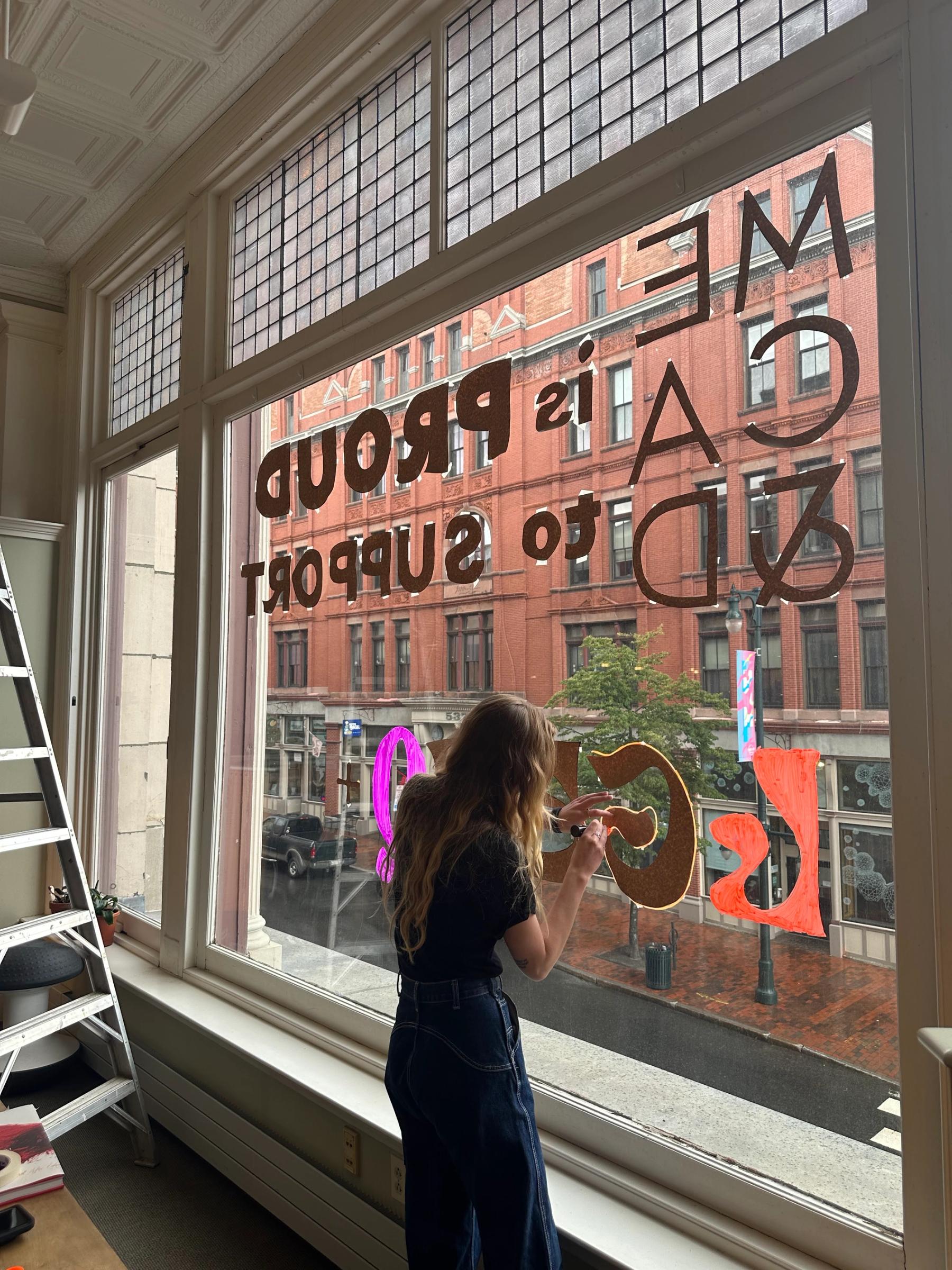 A woman stenciling "MECA&D is Proud to Support LGBTQ" on a large window.