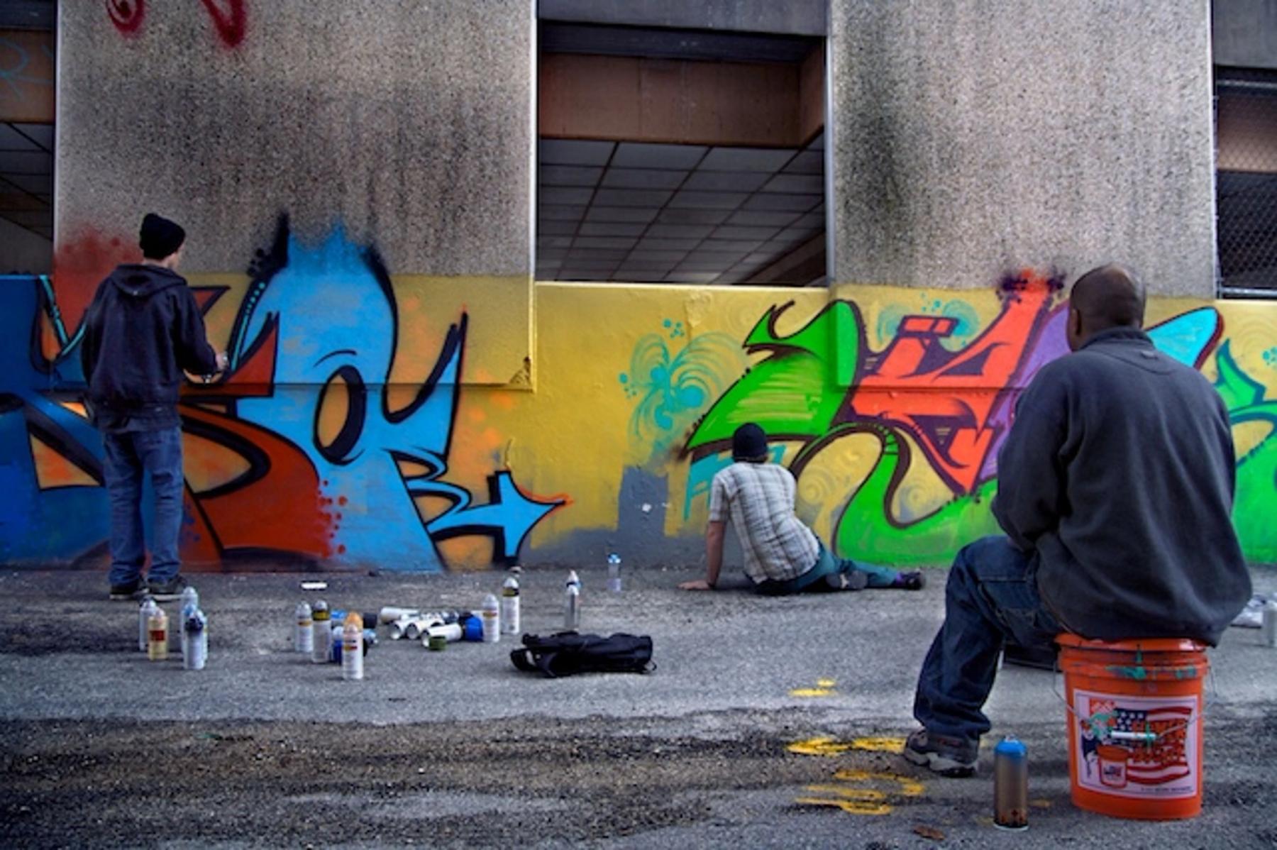 Two people spray painting a graffiti wall with one people sitting on a bucket, watching.