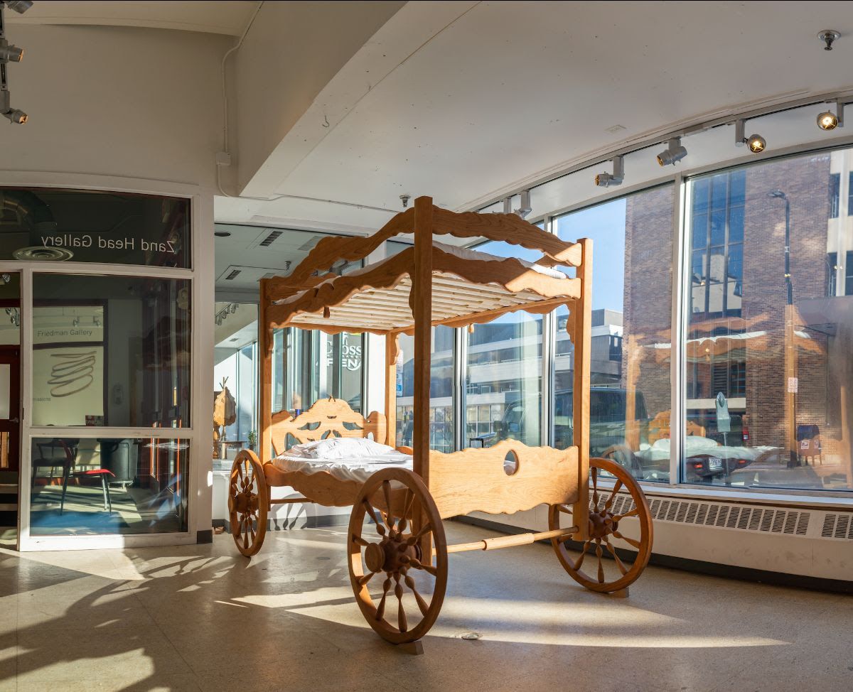 an art gallery with a wooden cart that has bunkbeds attached by posts to each wheel