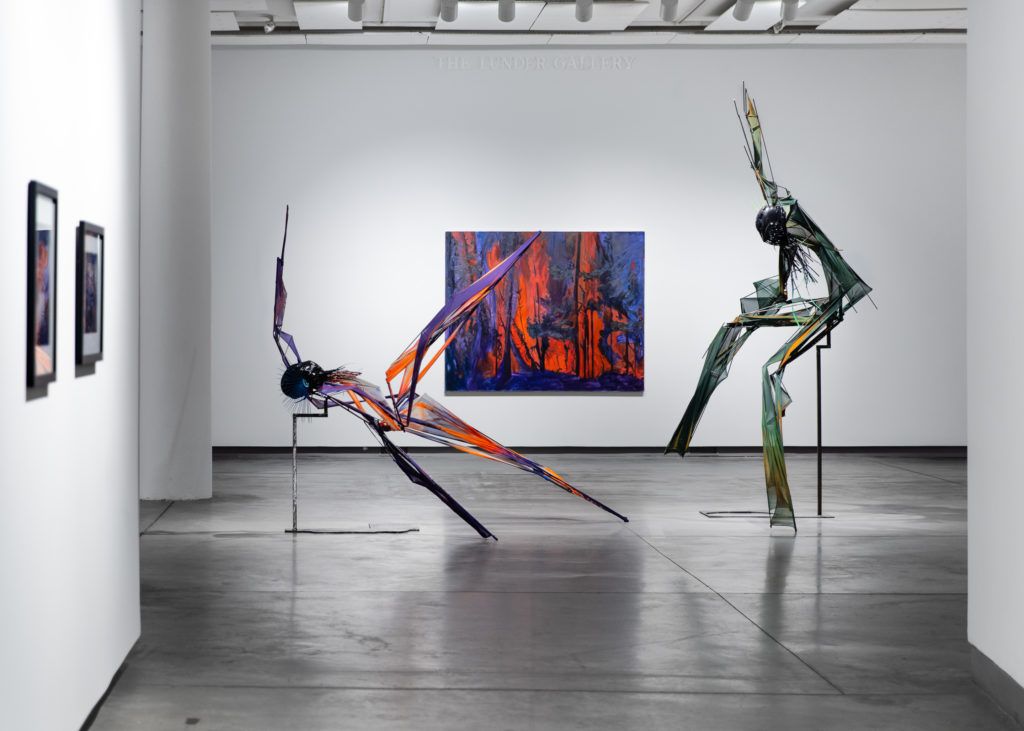Gallery view of two dynamic sculptures depicting dancing figures in front of a blue and red painting depicting burning trees.