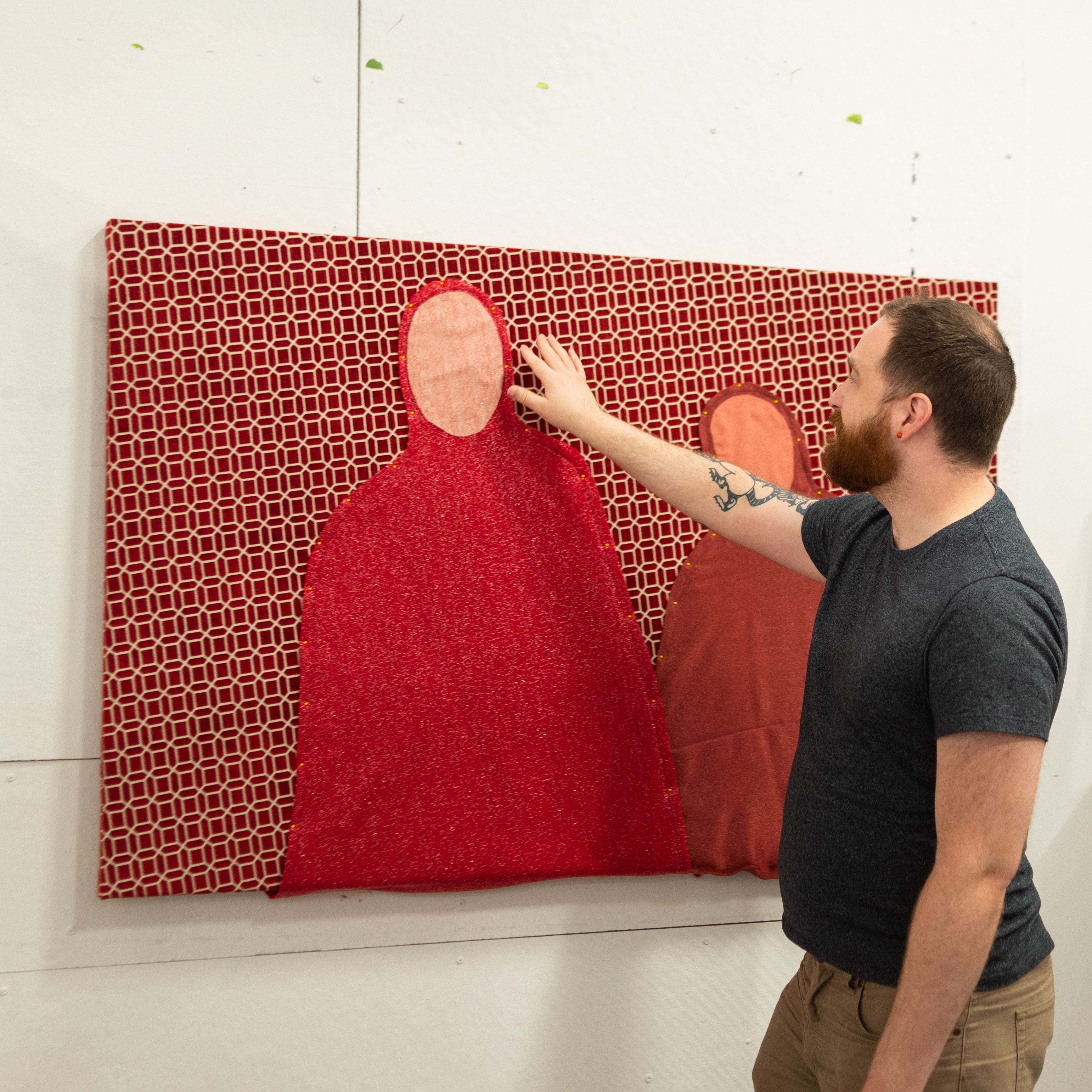 MFA student pointing at a fabric collage of two red figures over a patterned red background.