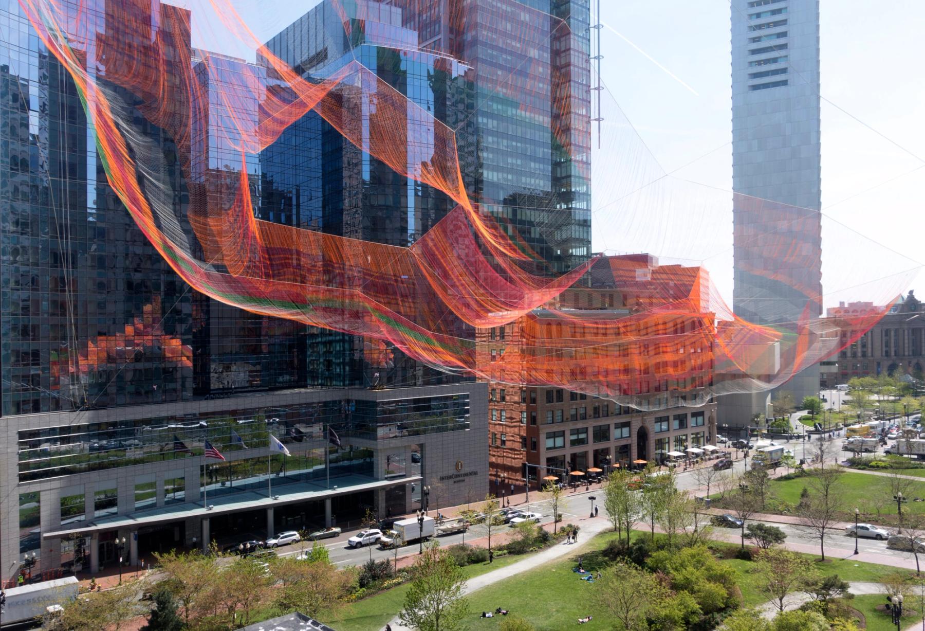 A massive red wave-like structure made of fibers against several tall buildings, above a park.