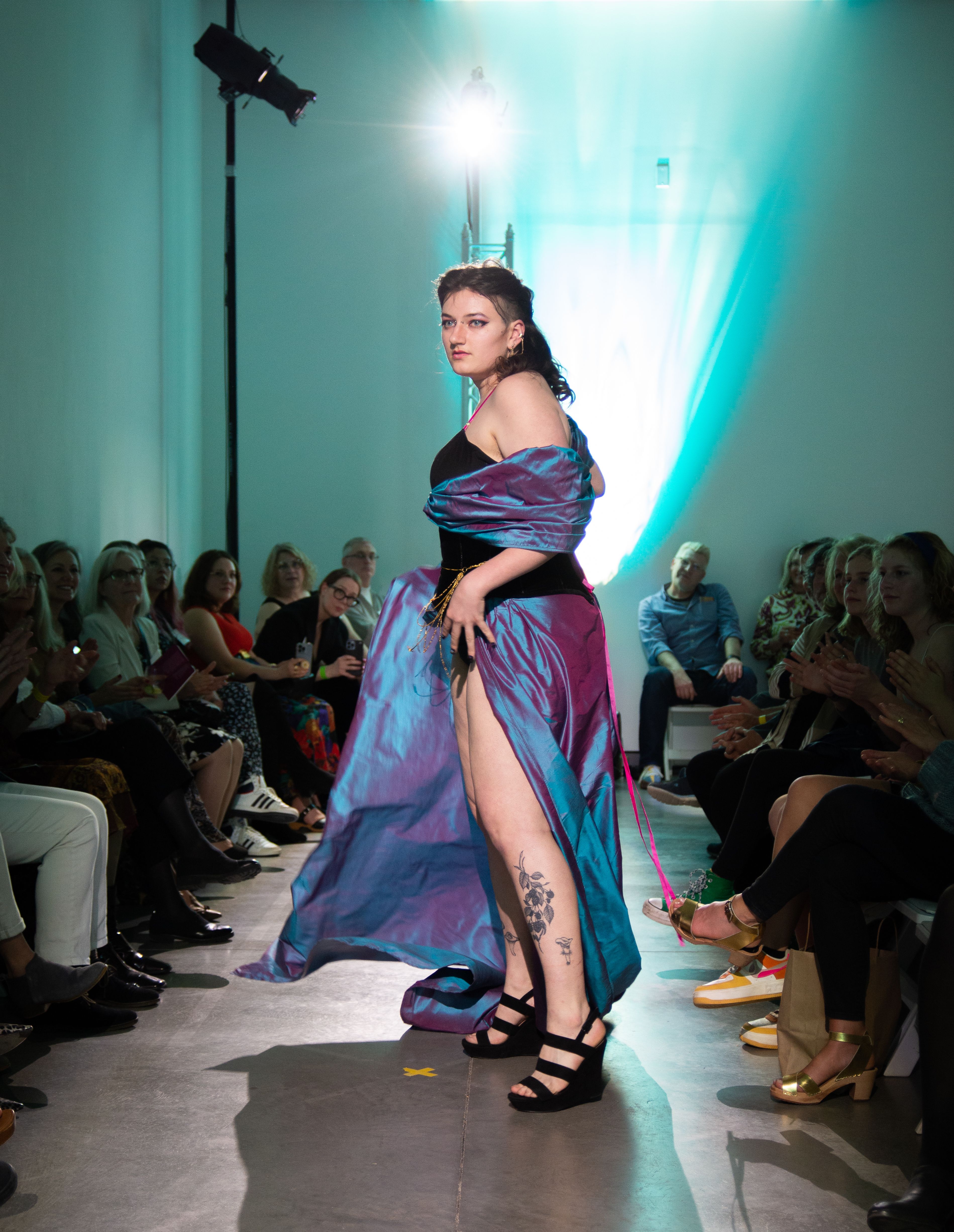 A model on the runway wearing gown with iridescent purple draped sleeves and skirt and a black bodice.