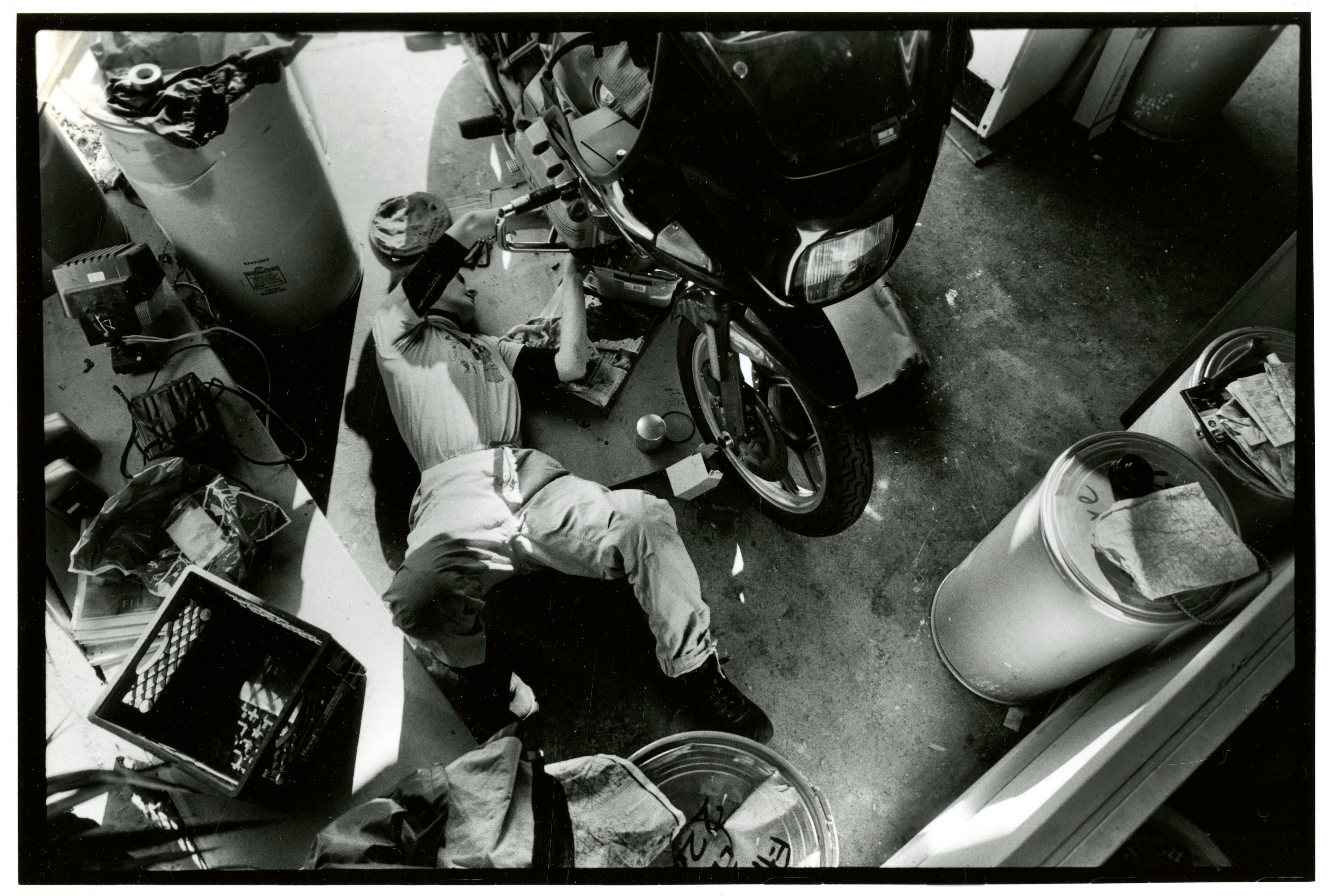 Black and white photograph of a mechanic laying on a garage floor to work on a motorcycle.