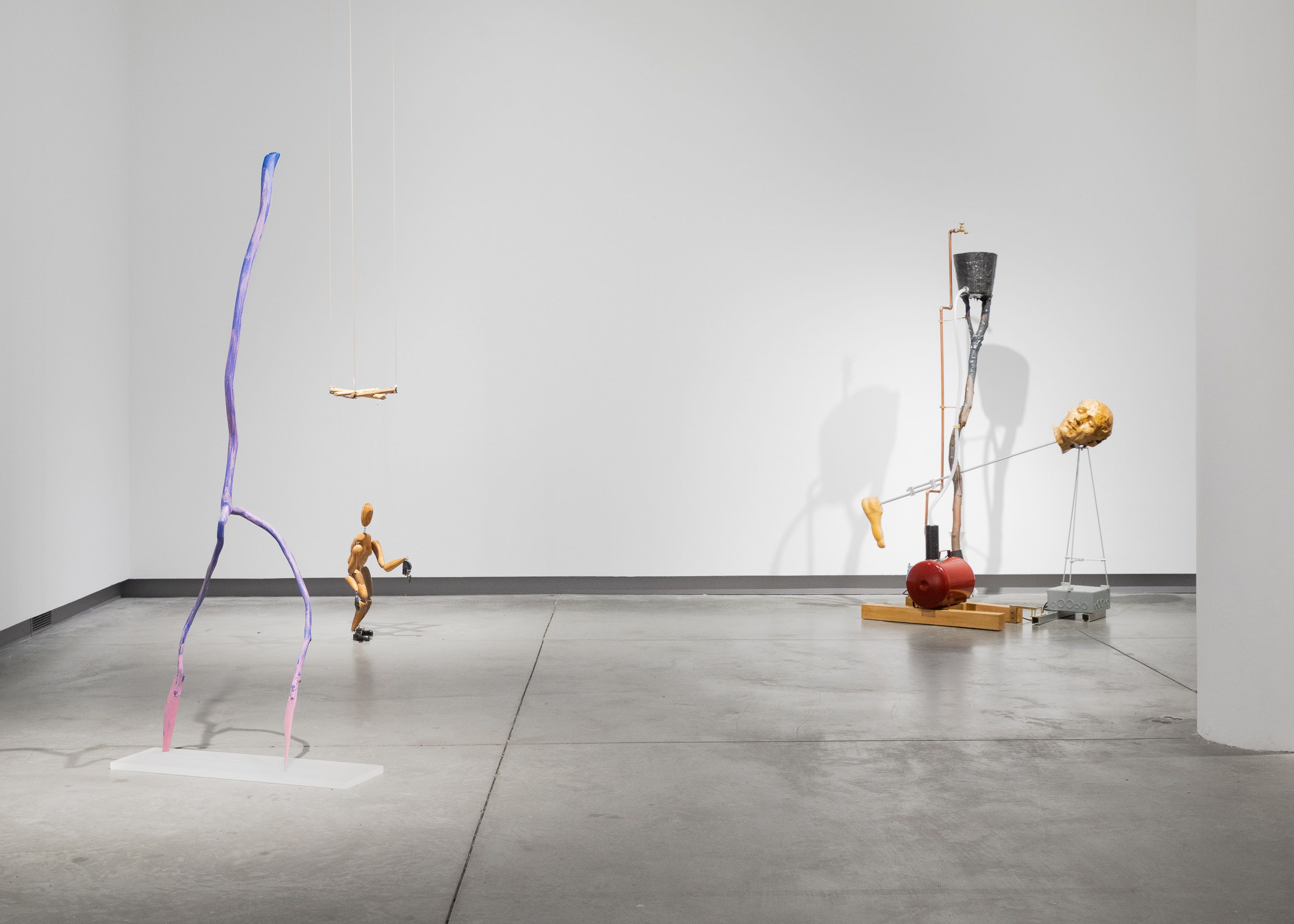 Installation view of three sculptures: the left a purple wire figure, the middle a marionette, and the left a kinetic foot and head.