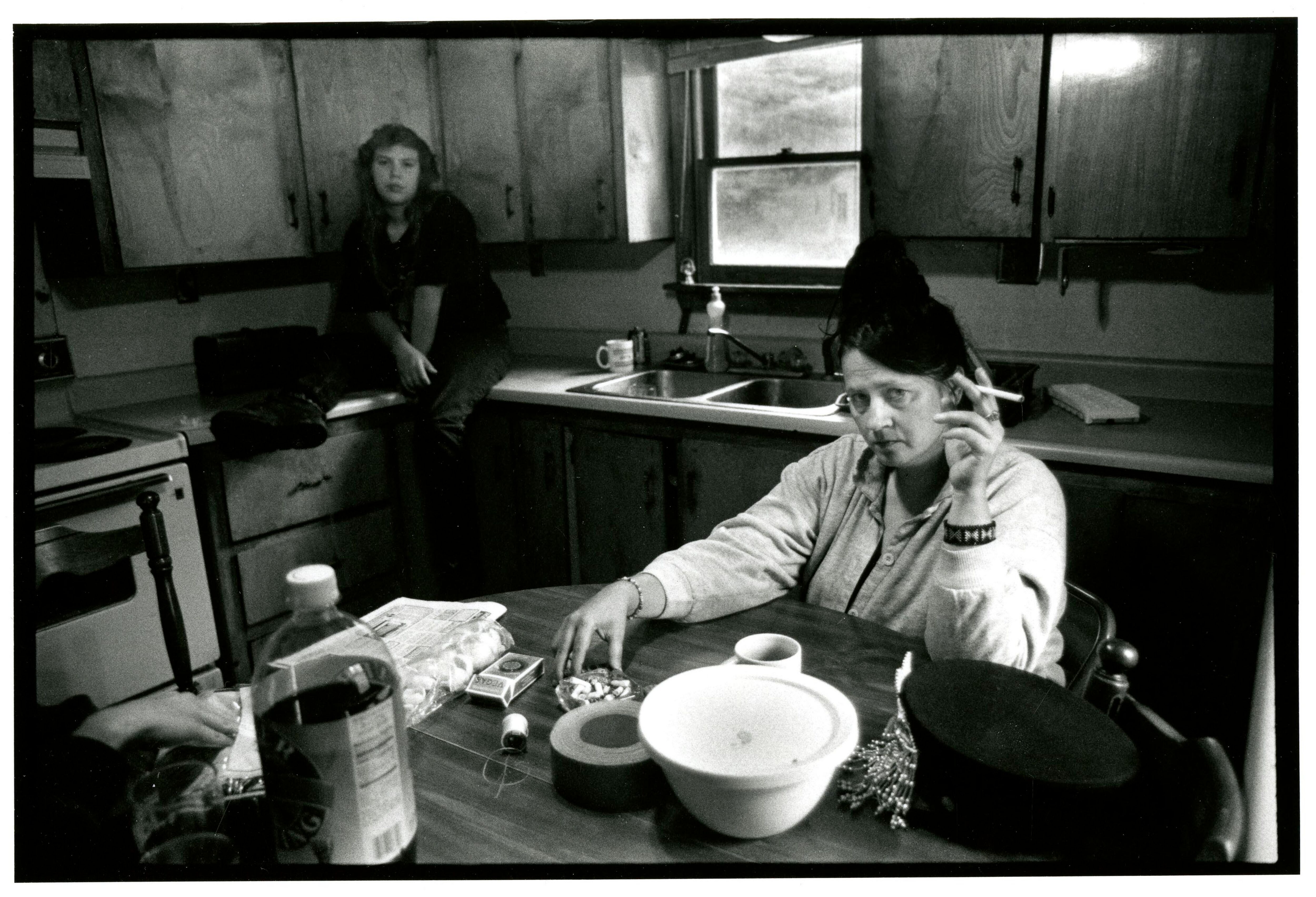 Black and white photograph of two women in a kitchen, the older sits at the table holding a cigarette, the younger sits on the counter.
