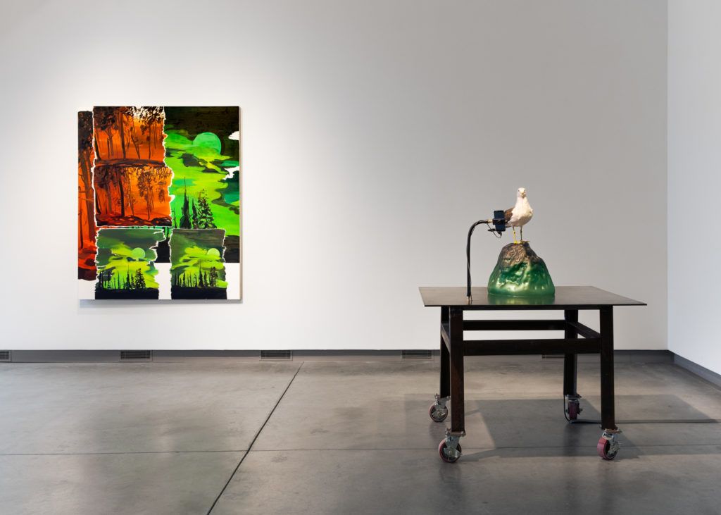 Gallery view of a red and green dystopian painting next to a sculpture depicting a seagull watching a phone.