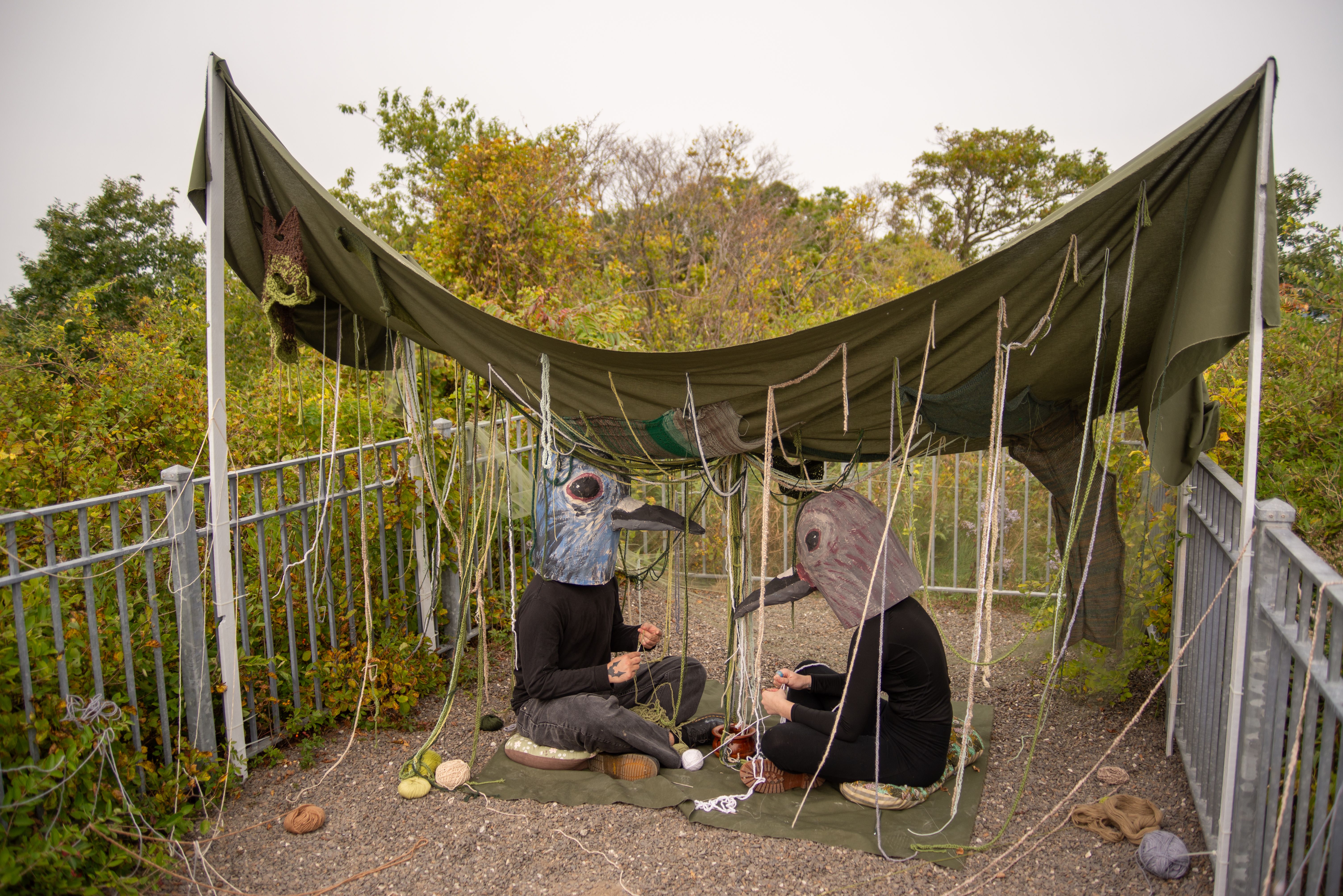 Two people sitting, facing each other, wearing large bird masks beneath a green overhang.