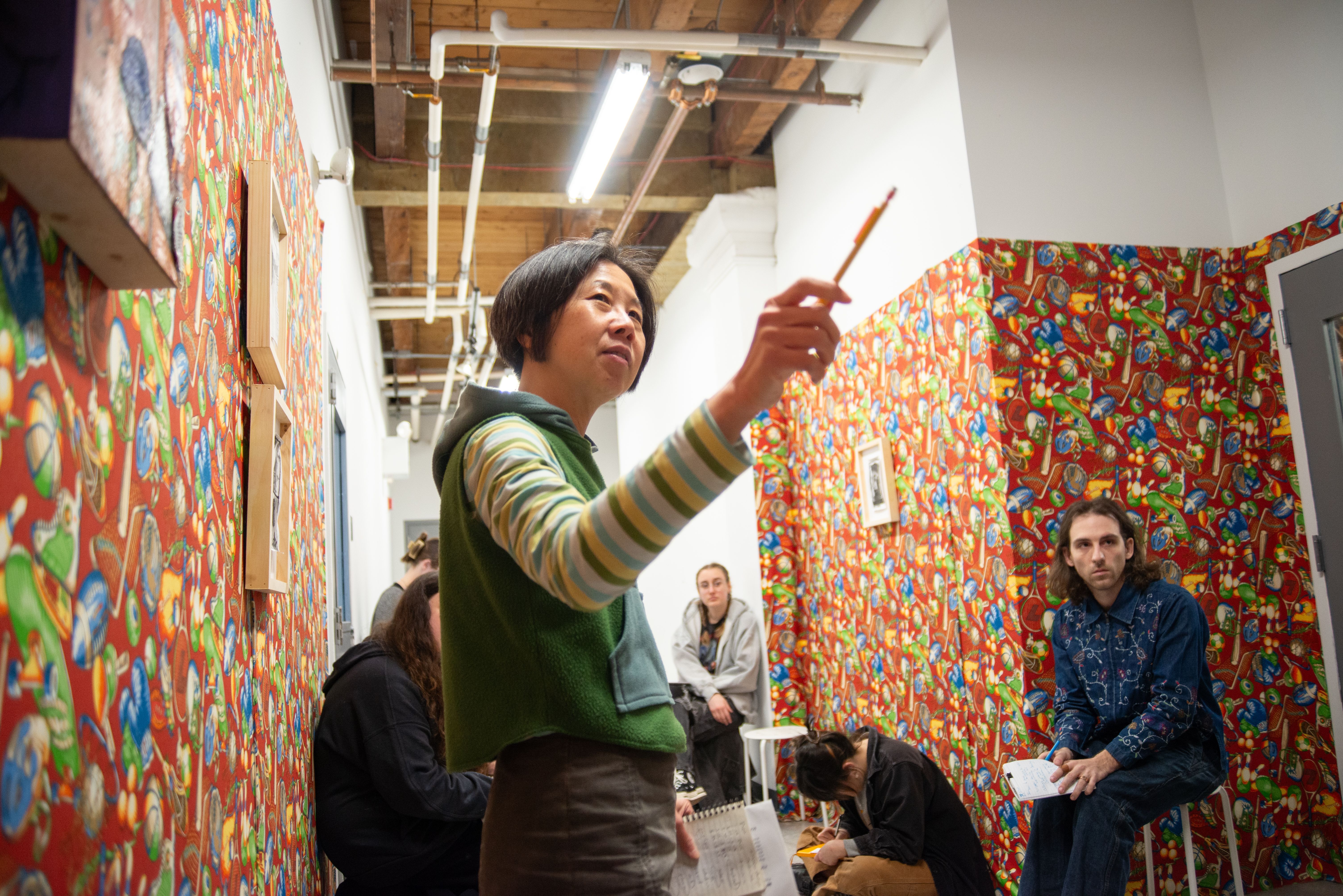 MFA critique with a professor pointing in a hallway covered with red wallpaper of sports balls.