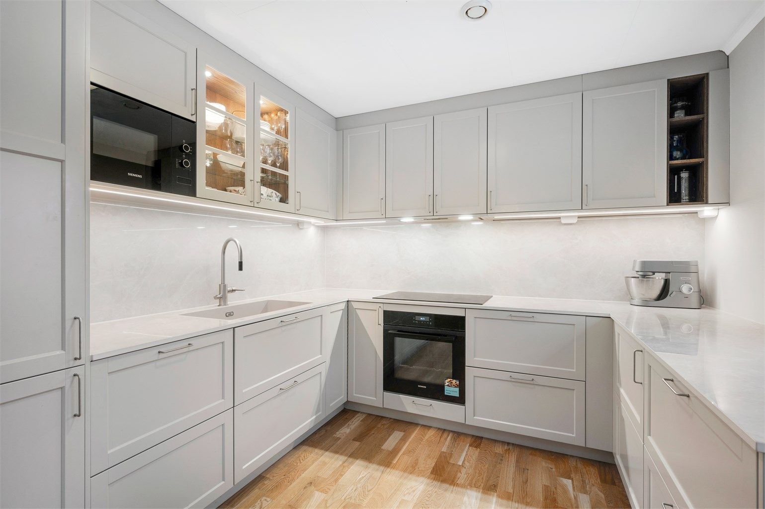 Nydelig kjøkkeninnredning fra Drømmekjøkkenet med integrert stekeovn, platetopp, oppvaskmaskin, mikrobølgeovn og kjøleskap med fryser. Legg merke til praktisk benkeskapsbelysning. 