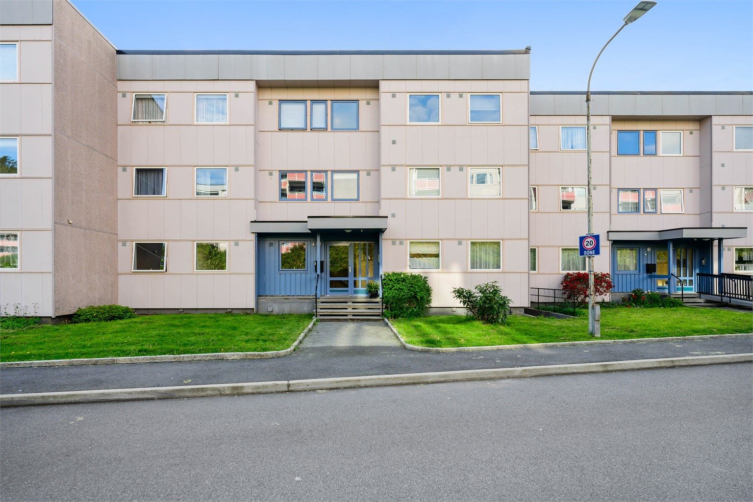 Inngangspartiet har grønne plener, beplanting og asfaltert gangvei.