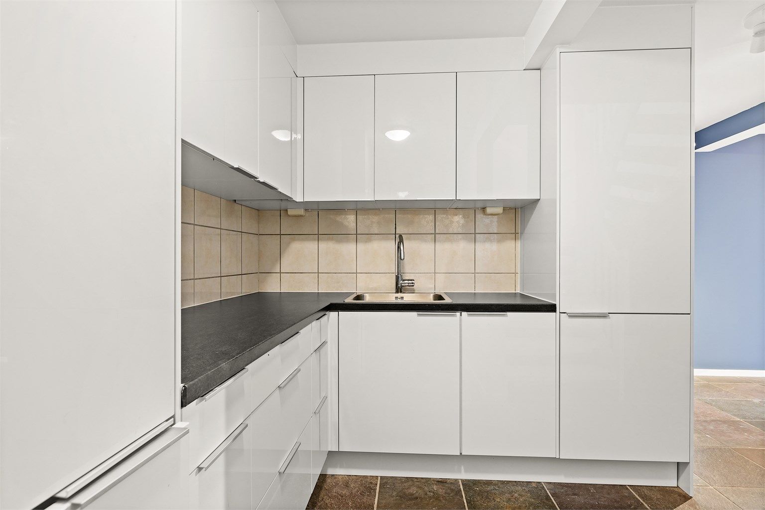 Kjøkkenet har hvite glatte fronter og laminert benkeplate, flislagt vegg mellom benkeplate og overskap.