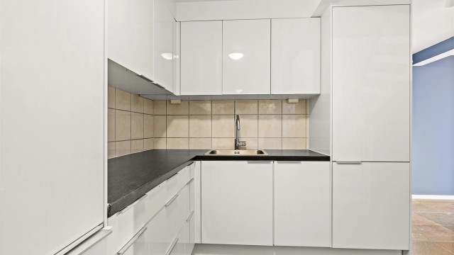 Kjøkkenet har hvite glatte fronter og laminert benkeplate, flislagt vegg mellom benkeplate og overskap.