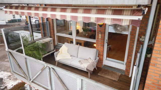 Balkongen er utstyrt med levegger av glass på rekkverk, utvendig belysning, strømuttak, markise, terrassevarmer og terrassebord på gulv.