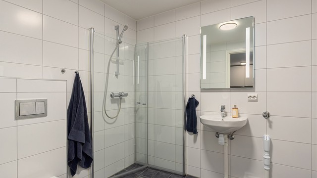 Lyst flislagt bad innredet med servant, toalett og dusjhjørne med innfellbare glassdører, samt opplegg for vaskemaskin.