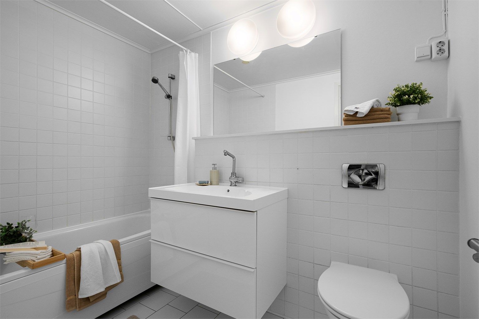 Bad I med fliser på gulv og delvis på vegg, varme i gulv. 
Det er vegghengt toalett, badekar med dusj, servant med skuffer .