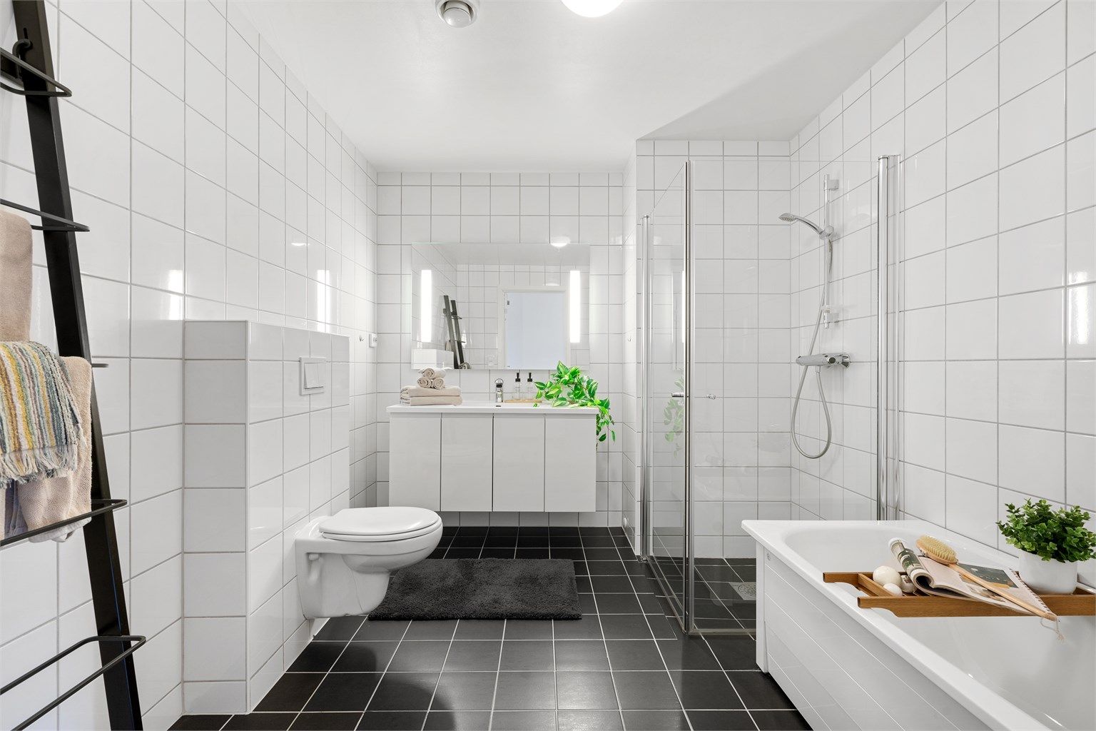 
Bad II er av lik design som bad I og har dusj med innfellbare dører, badekar, vegghengt toalett og servant med oppbevaringsplass.. Stort speil med innfelt lys på vegg. Det er mørke fliser på gulv og lyse på vegg. 