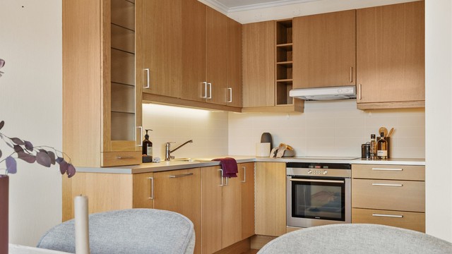 Åpen kjøkkenløsning. Innredning fra byggeår. Glatte fronter. Benkeplate med laminert overflate. Kjøkkenvask med armatur. Stikkontakt over kjøkkenbenk. Integrert stekeovn, platetopp, oppvaskmaskin og kjøleskap med fryser.