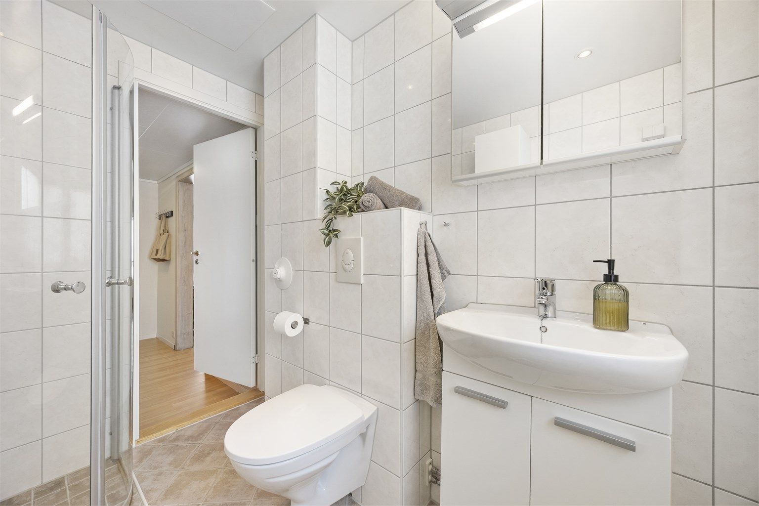 Badet er utstyrt med speilskap med overlys og stikkontakt over servant, dusjhjørne med glassdører og veggmontert dusjarmatur samt vegghengt toalett med innebygget sisterne.