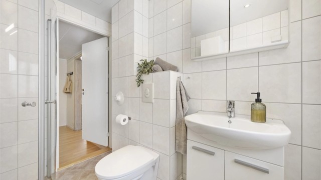 Badet er utstyrt med speilskap med overlys og stikkontakt over servant, dusjhjørne med glassdører og veggmontert dusjarmatur samt vegghengt toalett med innebygget sisterne.