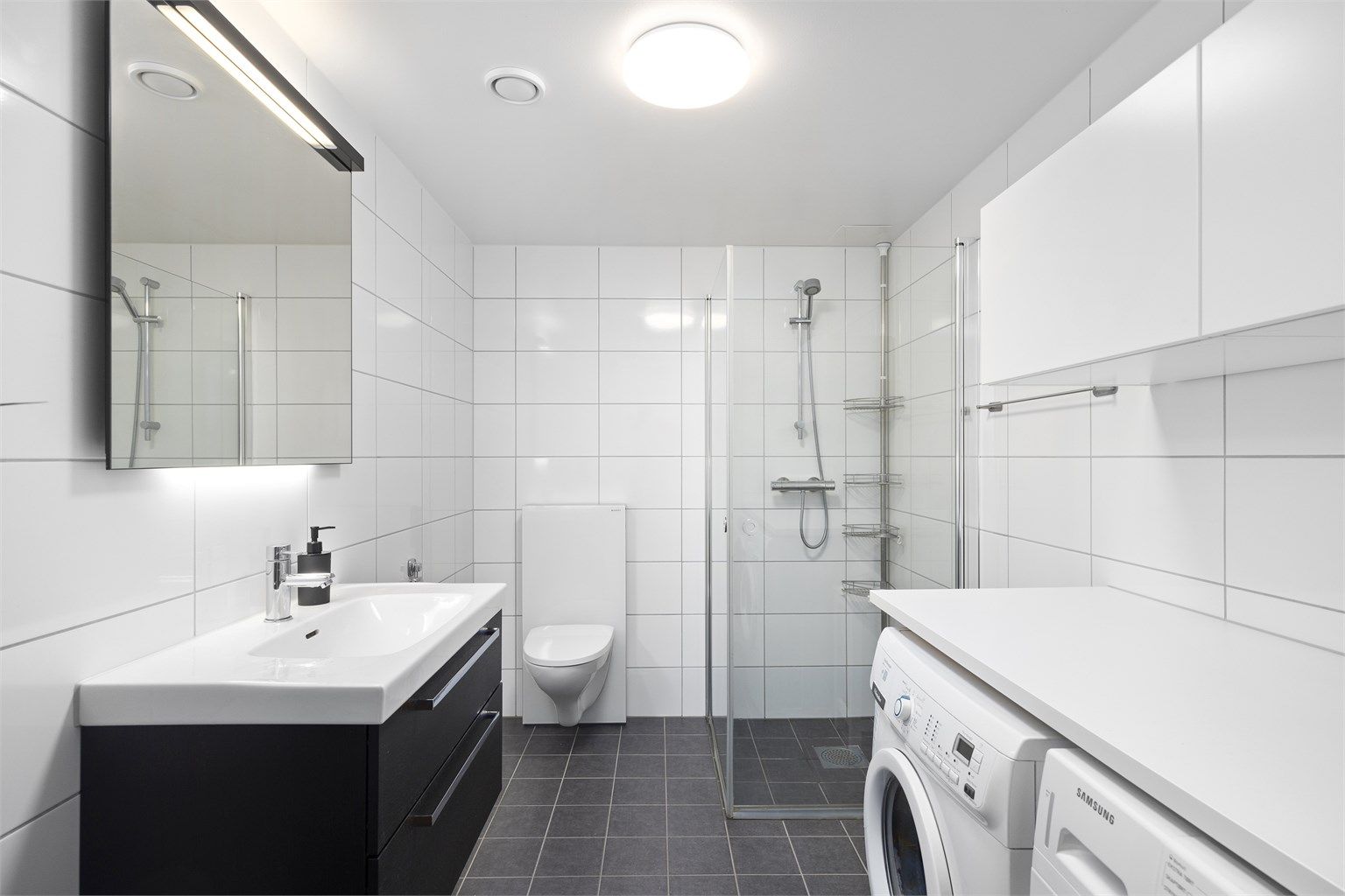 Innbydende og meget romslig bad med hvite fliser på vegg og grå fliser med varme i gulv