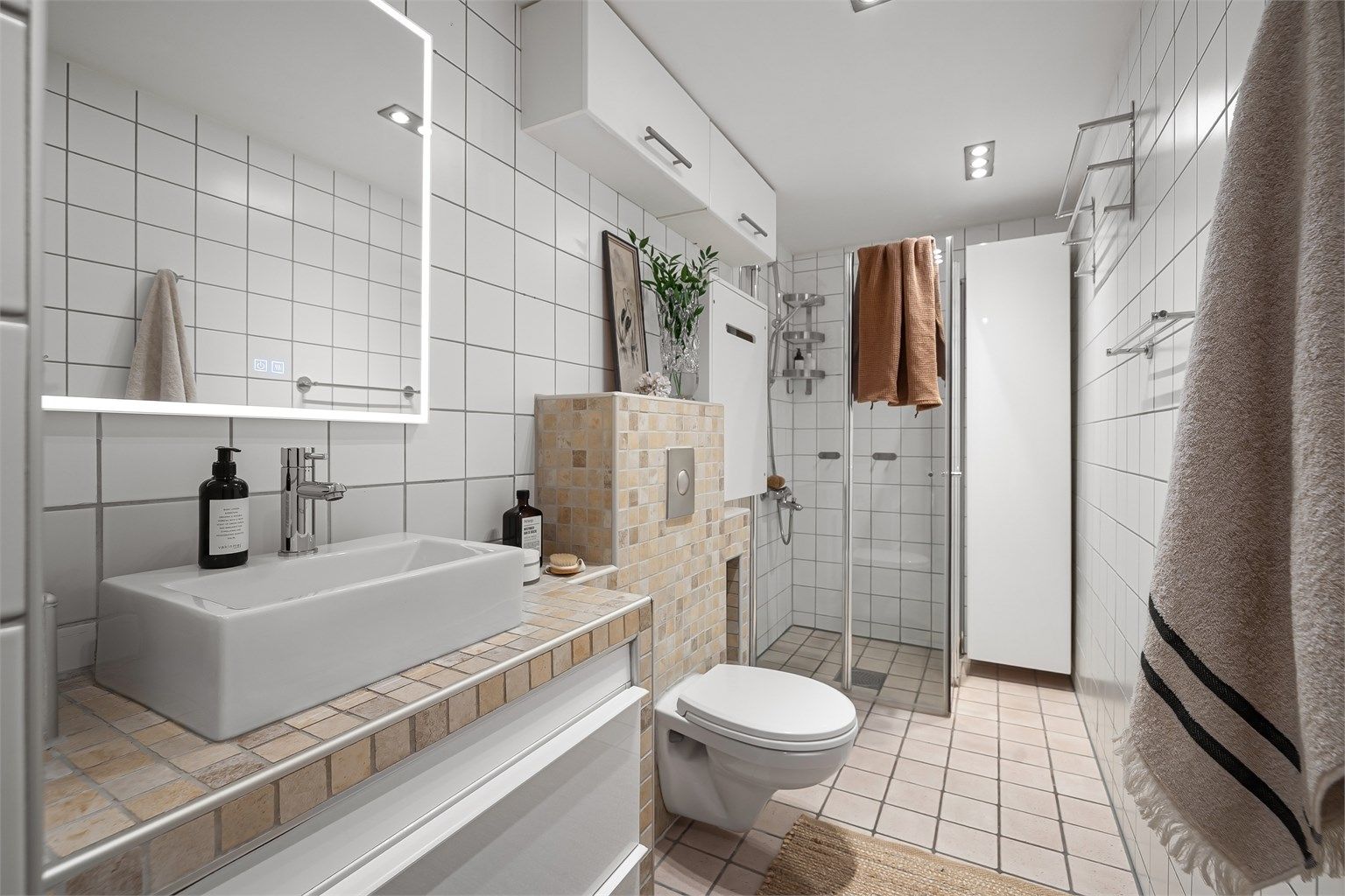 Badet er utstyrt med vegghengt toalett og dusjhjørne med innfellbare glassdører, hvorav én dør er leddet