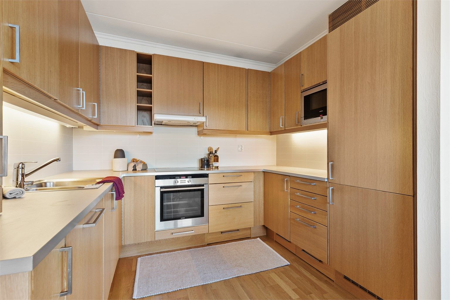 Åpen kjøkkenløsning. Innredning fra byggeår. Glatte fronter. Benkeplate med laminert overflate. Kjøkkenvask med armatur. Stikkontakt over kjøkkenbenk. Integrert stekeovn, platetopp, oppvaskmaskin og kjøleskap med fryser.