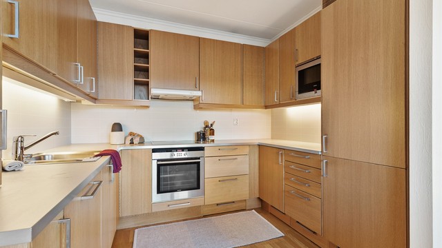 Åpen kjøkkenløsning. Innredning fra byggeår. Glatte fronter. Benkeplate med laminert overflate. Kjøkkenvask med armatur. Stikkontakt over kjøkkenbenk. Integrert stekeovn, platetopp, oppvaskmaskin og kjøleskap med fryser.