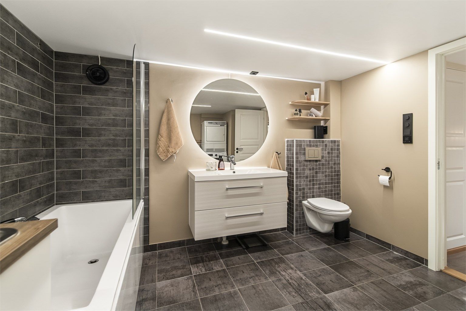 Romslig baderom/kombinert vaskerom i kjelleren fra 2017. Det er fliser på gulv med varme, og veggflatene består av malte, slette flater samt noe fliser ved badekaret. Himlingen er malt og har stilige, innfelte LED-lys. 