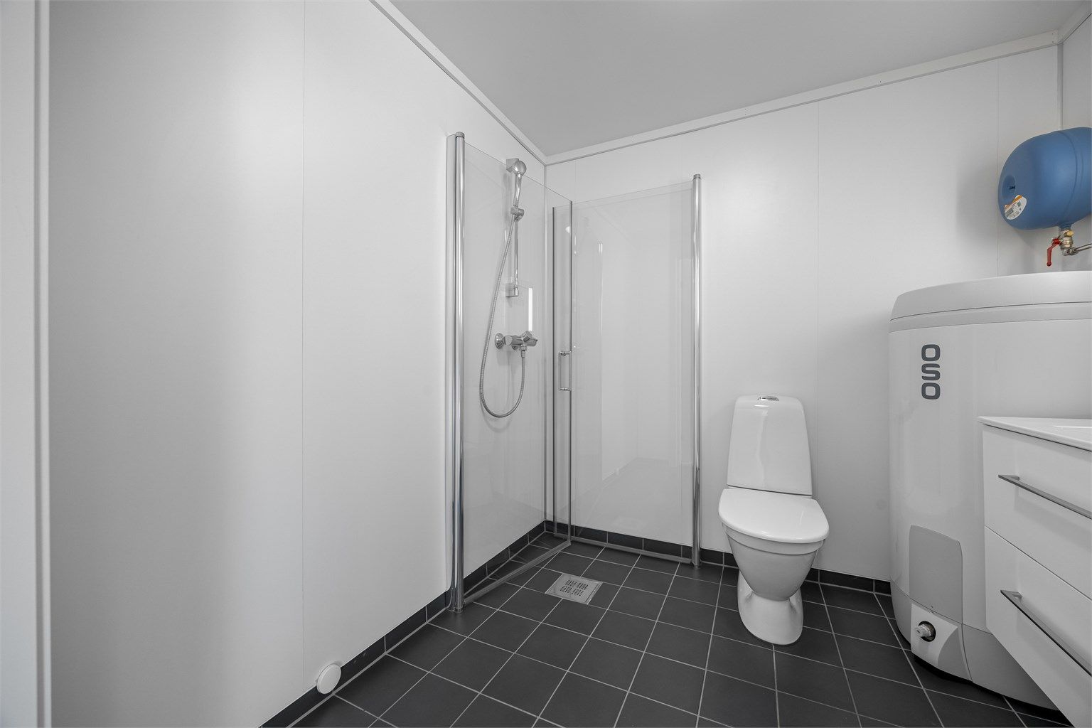 Baderom med opplegg til vaskemaskin, dusjhjørne, toalett, varmtvannsbereder (200l) og servant med speil og skuffer.