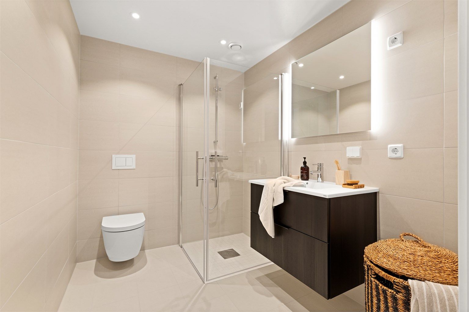 Badet har en ren og moderne utforming med store, sandfargede fliser og vannbåren gulvvarme.