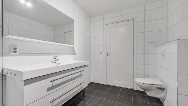 Baderom fra byggeåret med servant i innredningen, speil og vegghengt toalett, samt dusjhjørne med glassvegger