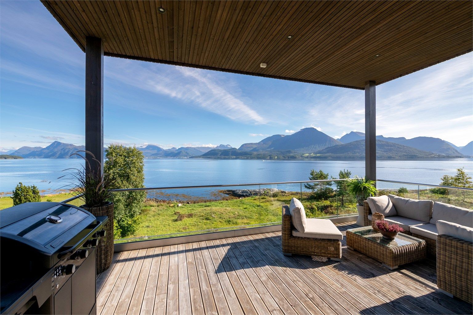 Overbygd balkong med glassrekkverk, terrassebord og utsikt mot fjordlandskapet.