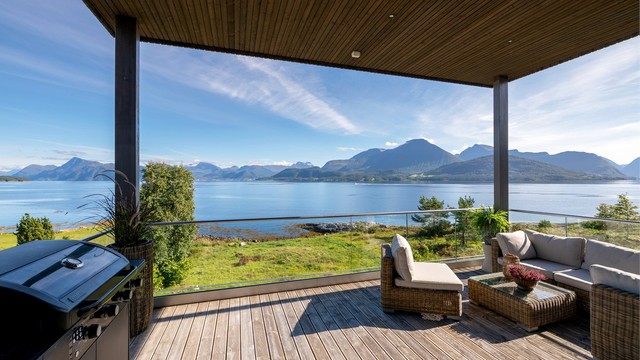 Overbygd balkong med glassrekkverk, terrassebord og utsikt mot fjordlandskapet.