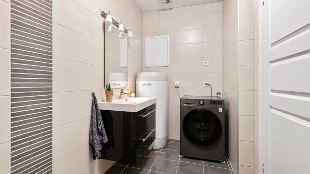 Sort servantinnrending med skuffer og glatte fronter i høyglans. Ovenpåliggende servant med ett-greps armatur. Speil med overlys. Opplegg for vaskemaskin. Varmtvannsbereder plassert i hjørne