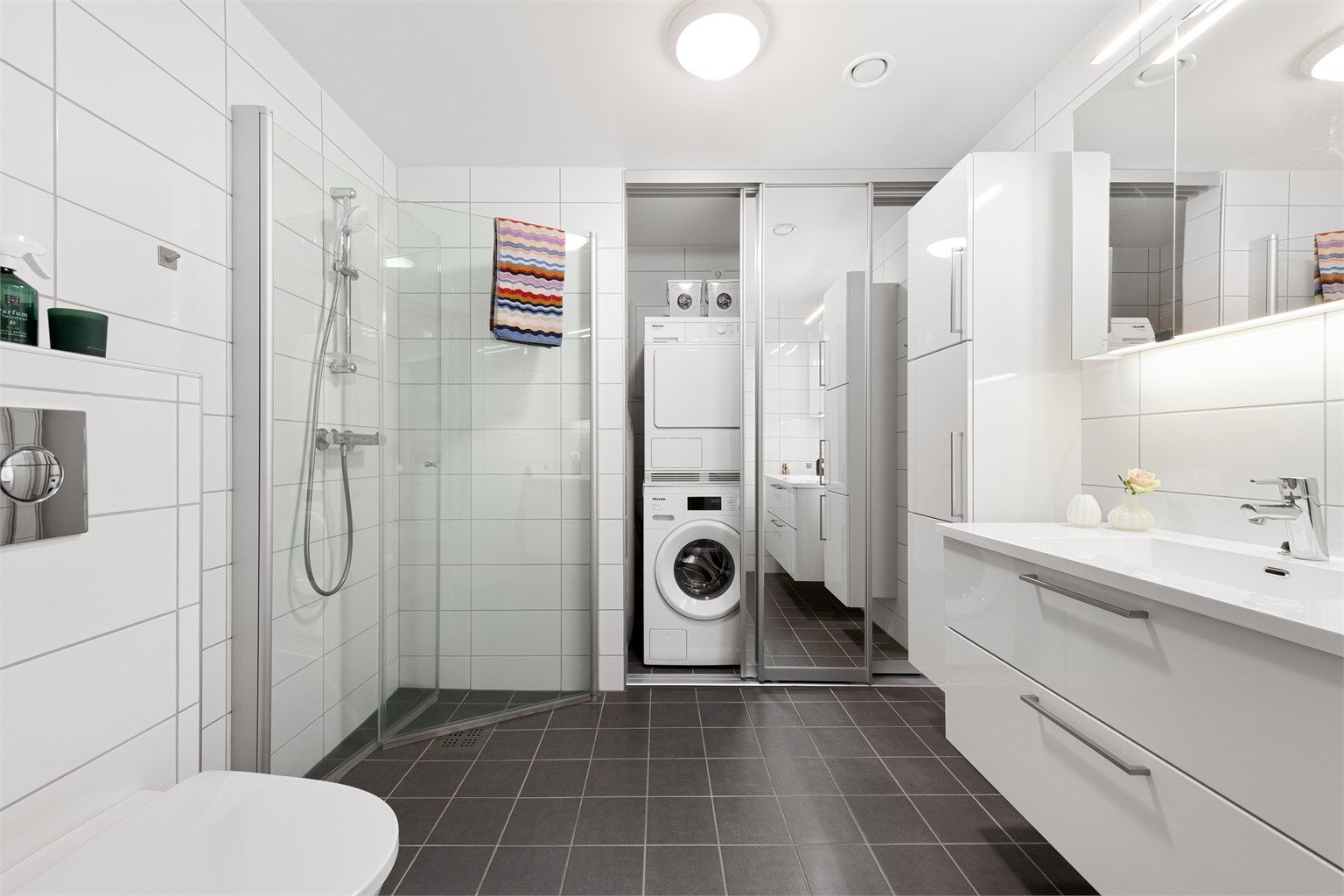 Flislagt bad med varmekabler i gulv. Det er skyvedørsfronter med speildører som skjuler varmtvannsbereder, stoppekran, opplegg for vaskemaskin og ventilasjonsaggregat.