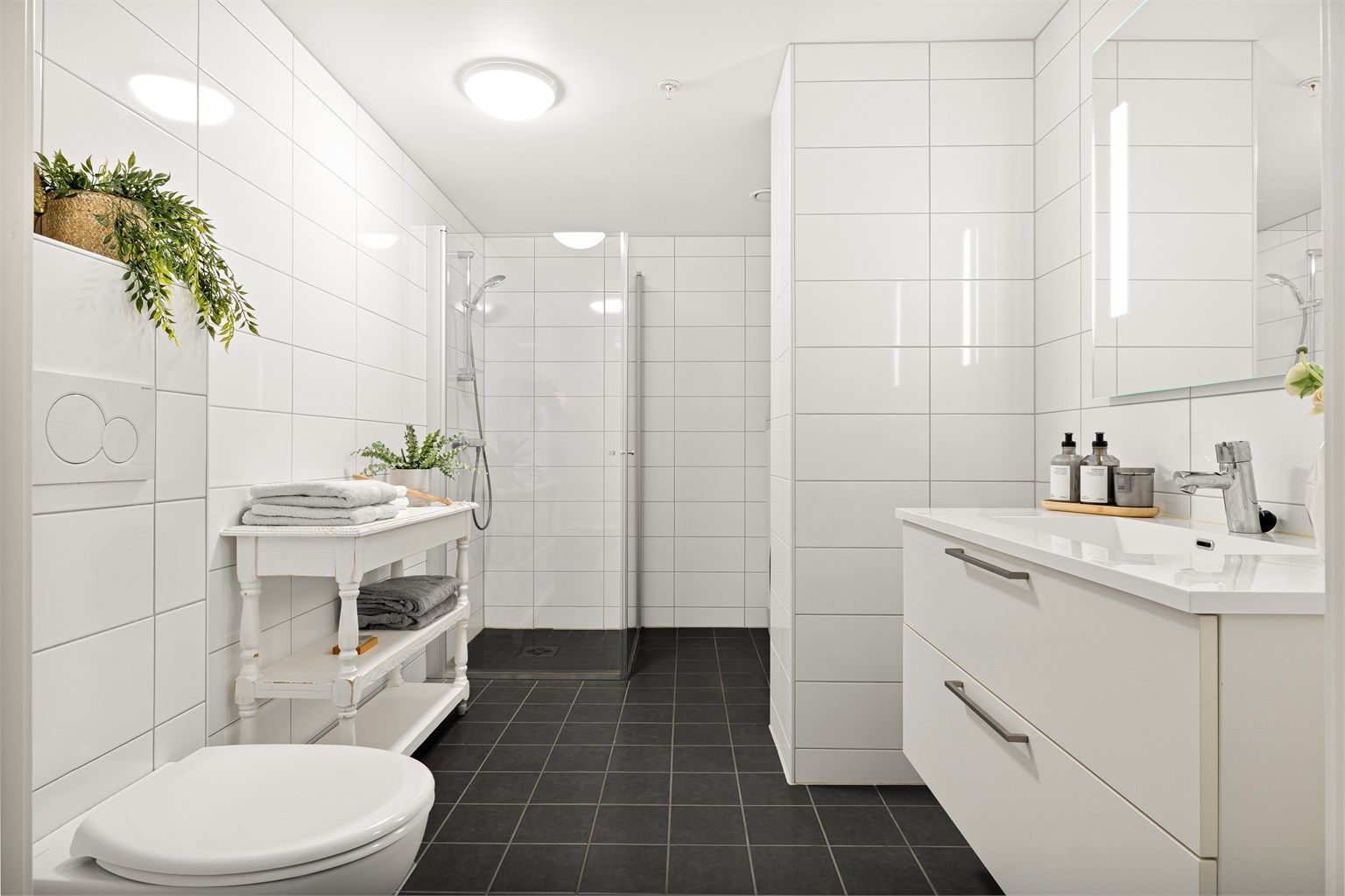 Tidløst og helfliset bad med varme i gulv. Badet er innredet med vegghengt toalett, dusj med glass foldedører, servant med møblement, speil og belysning.